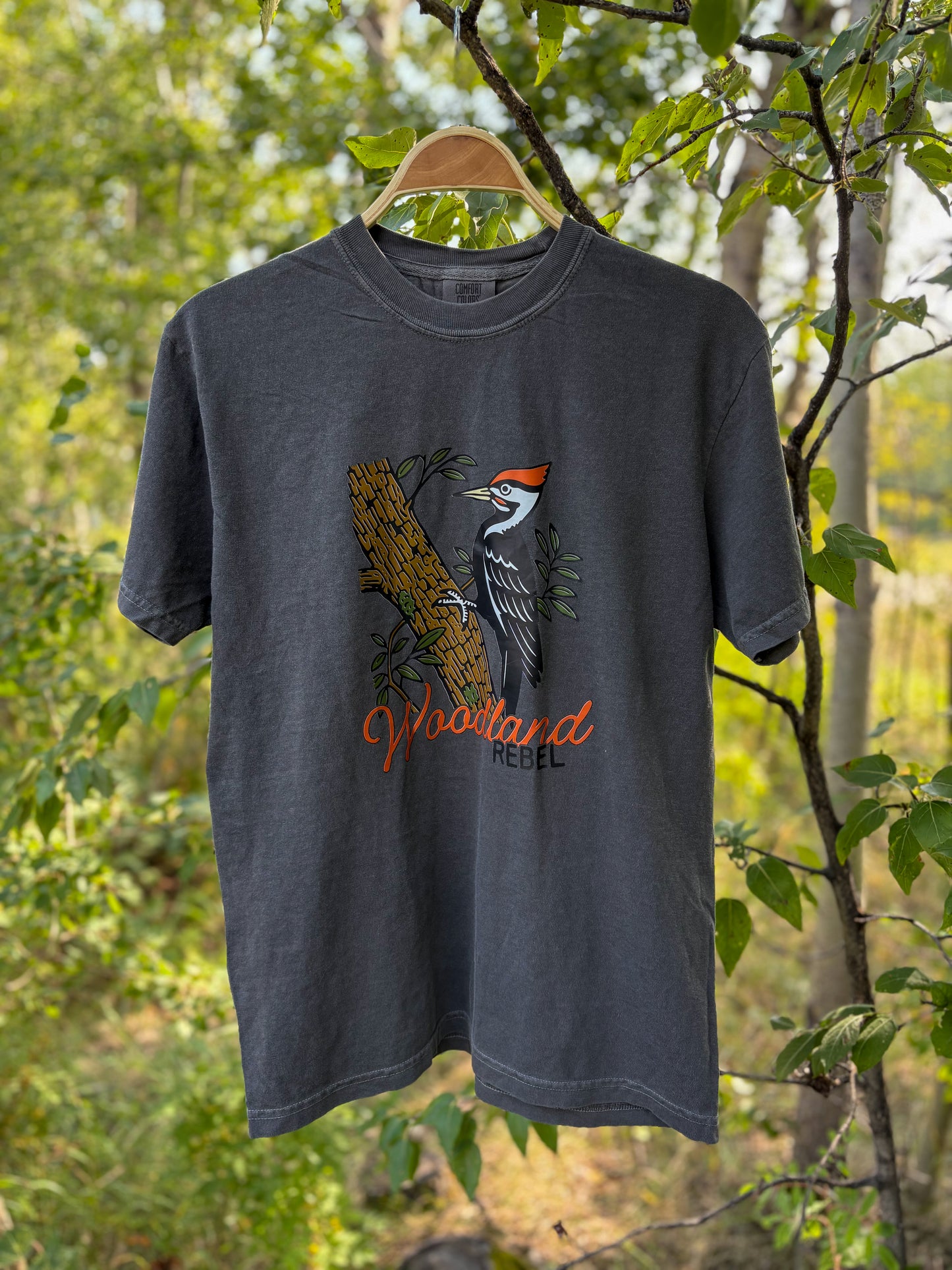 Gray t-shirt with a woodpecker design hanging on a tree branch in a forest setting