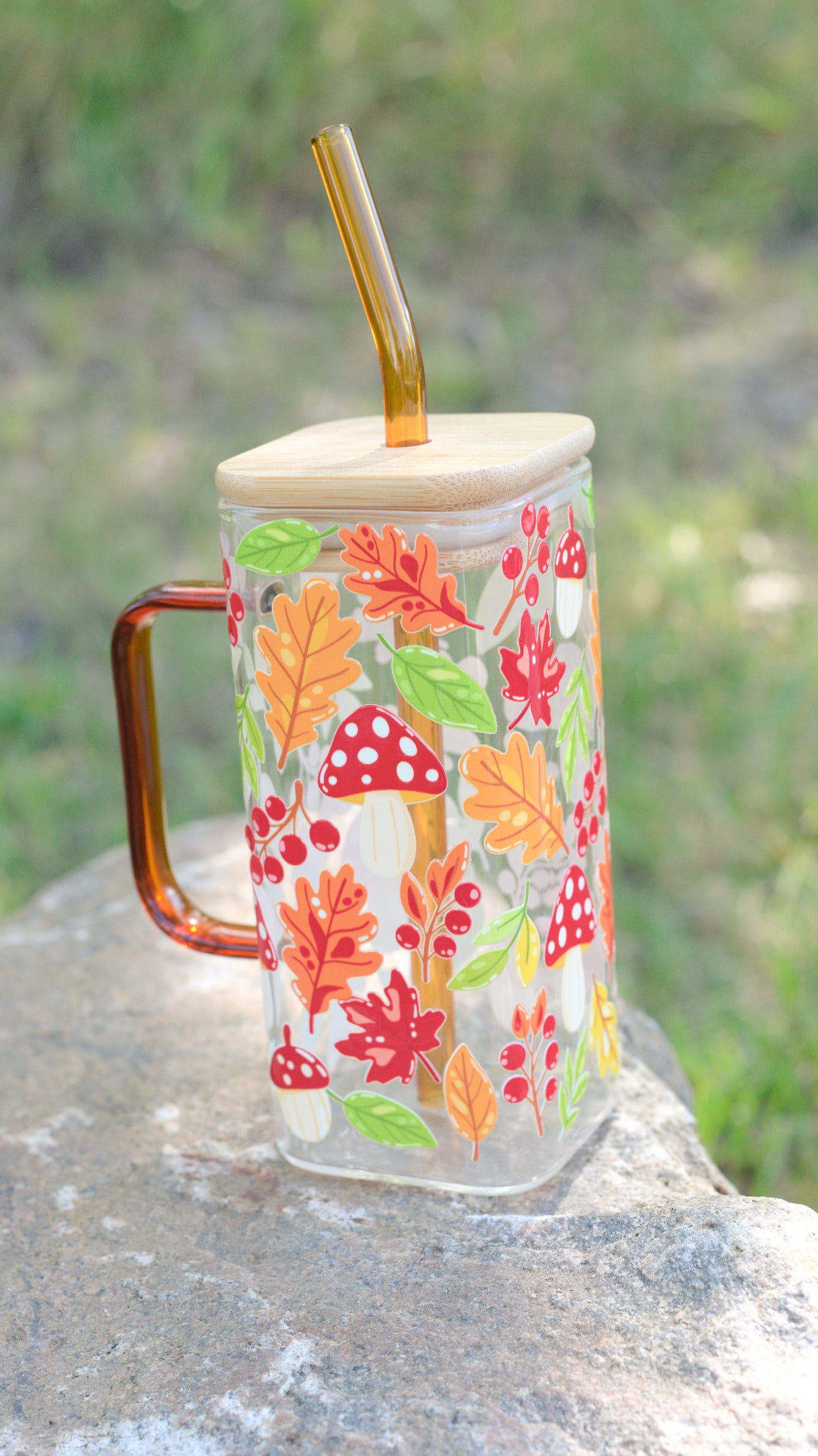 Glass square mug with autumn leaf and mushroom design, coloured glass handle, and straw on a natural background.