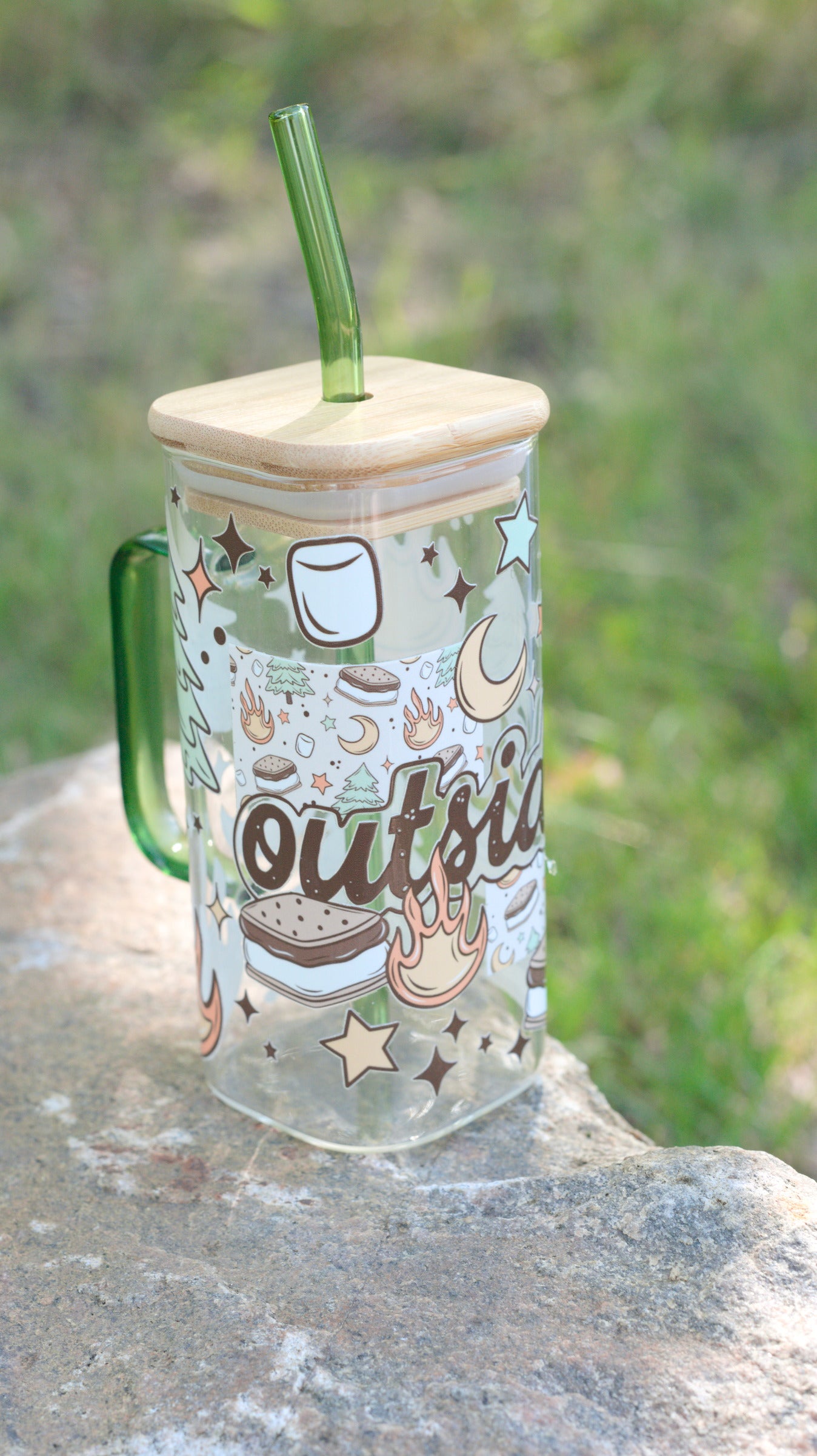 Clear mug with nature-themed design, wooden lid, and green straw on a stone surface with blurred greenery in the background.