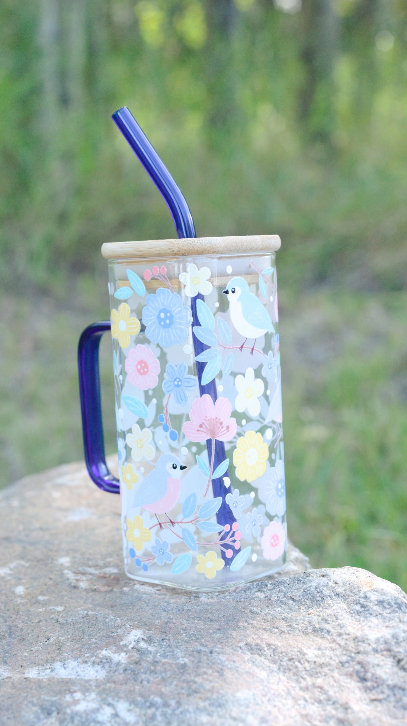 Glass mug with floral design, handle, and straw on a rock with greenery in the background