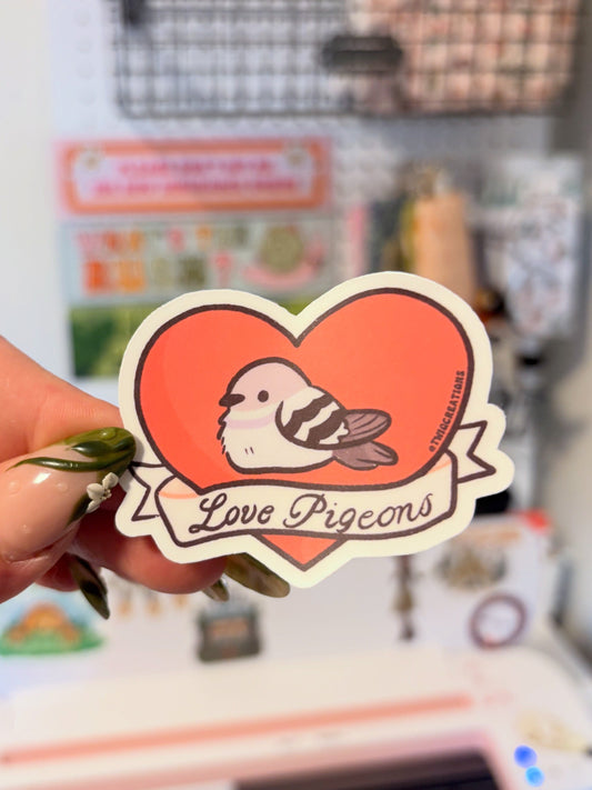 A hand with green nail polish holds a heart-shaped sticker featuring a pigeon bird on an orange-red heart with the words 'Love Pigeons' and a small 'PigeonArt' brand mark, against a blurred craft room background.